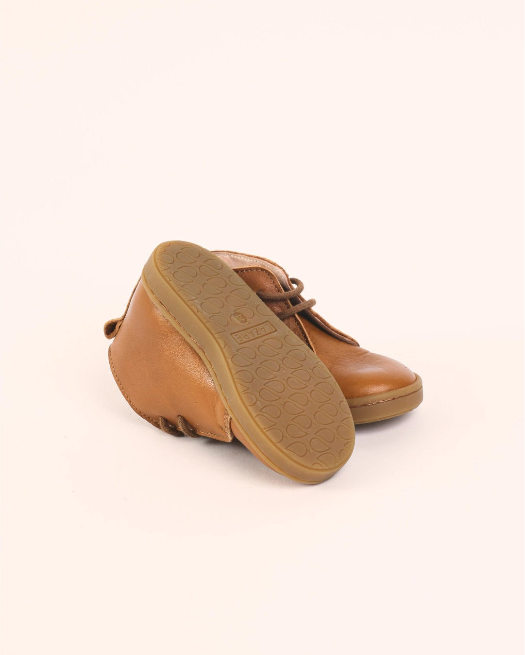 A pair of tan leather lace-up shoes shown from a high angle, with one shoe turned to display its brown textured sole, set against a solid pale peach background.