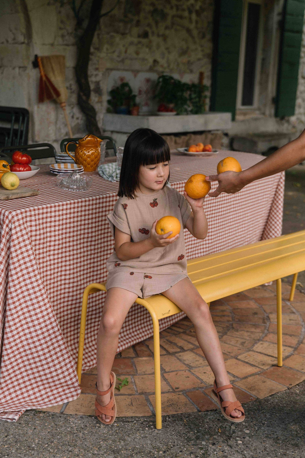 Child sitting on a yellow chair holding oranges in an outdoor setting with a table and bench.