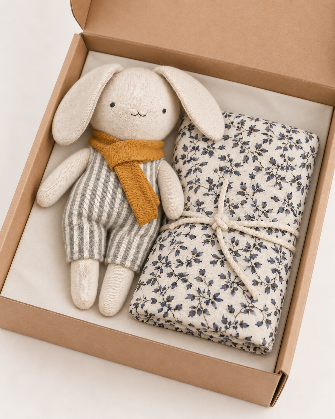 A plush bunny toy with long ears, cream-colored fabric, and a mustard yellow scarf, displayed next to a folded cream fabric item with a dark floral print inside an open brown cardboard gift box.