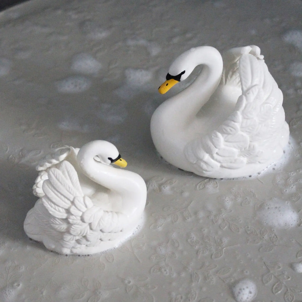 Two white swan figurines with yellow beaks and black eyes, placed on a textured gray surface with soap bubbles around them.