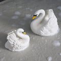 Two white swan figurines with yellow beaks and black eyes, placed on a textured gray surface with soap bubbles around them.