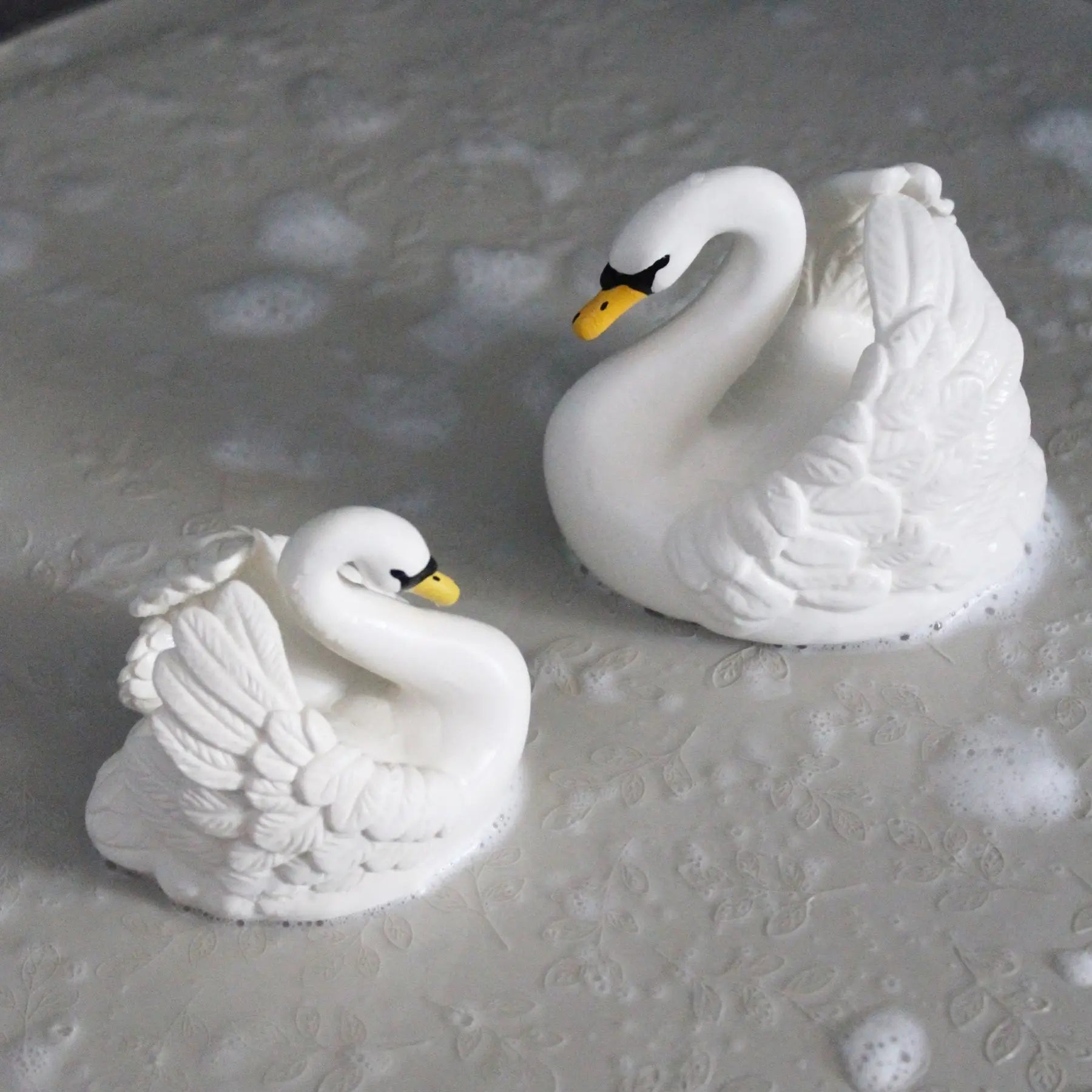Two white swan figurines with yellow beaks and black eyes, placed on a textured gray surface with soap bubbles around them.