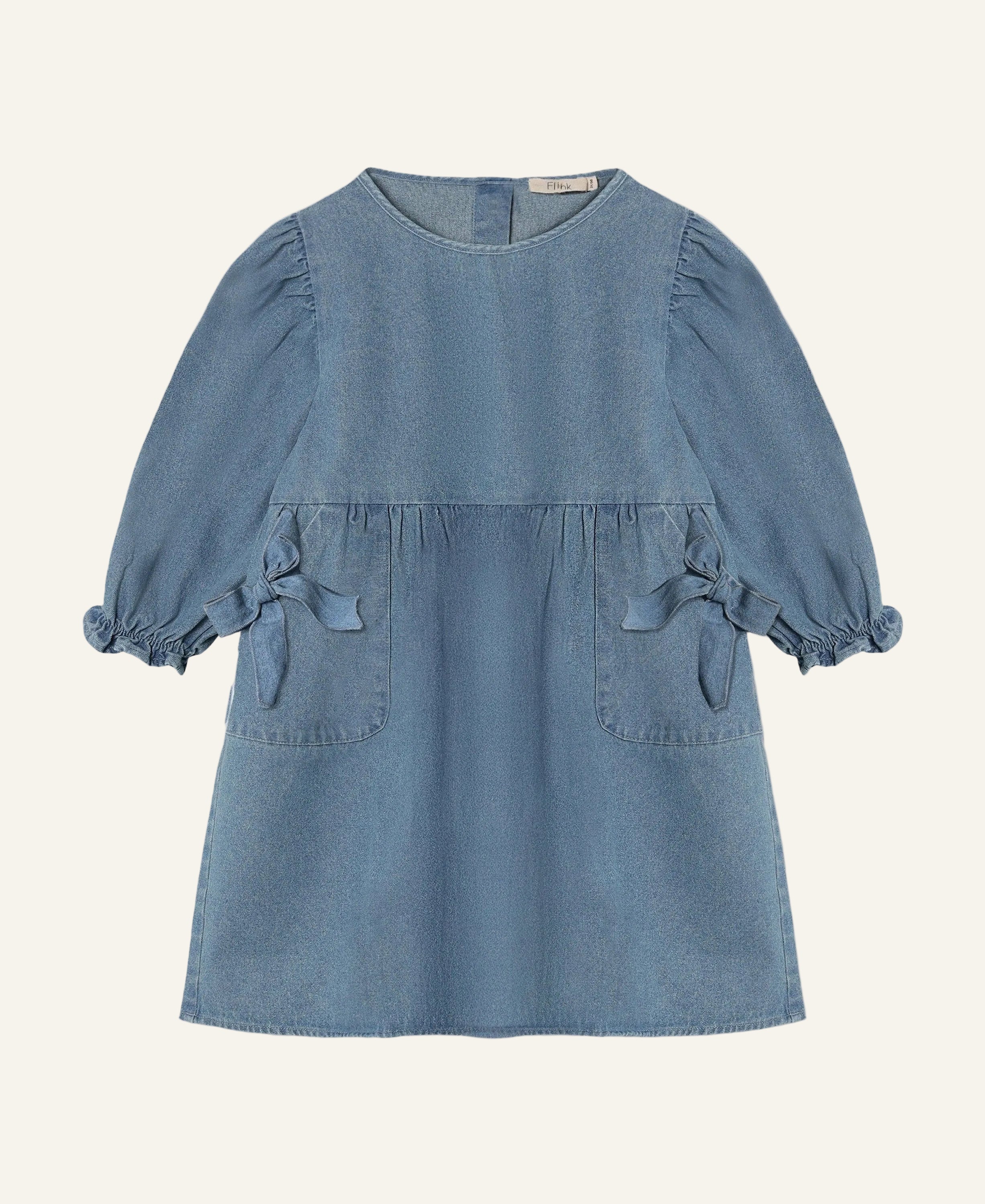 A light blue denim dress for toddlers, featuring a round neckline, gathered puff sleeves with tie details at the cuffs, and two front patch pockets, shown against a plain light gray background.