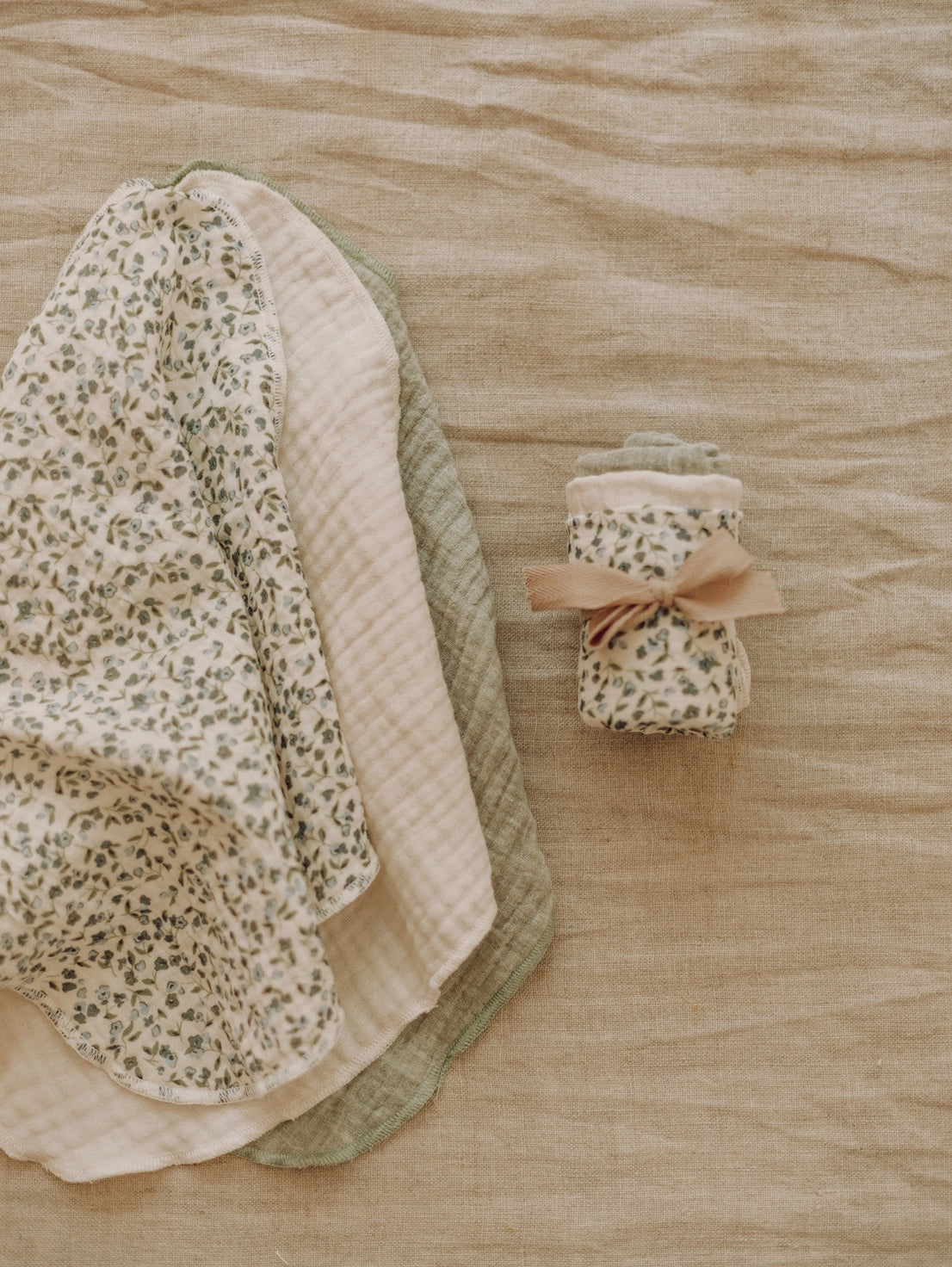 A set of baby muslin cloths and a matching rolled swaddle set on a beige linen background. The cloths feature a green floral print on cream, a solid pale pink/cream side, and a sage green quilted layer.