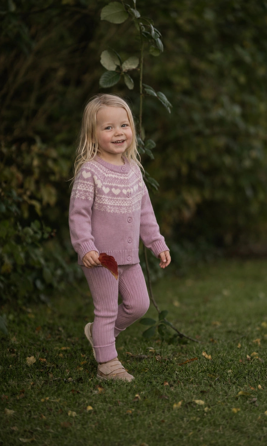 A young blonde girl wearing a matching dusty pink knit cardigan and matching pants stands on a grassy lawn with a leaf in her hand, set against a soft-focus background of green bushes.