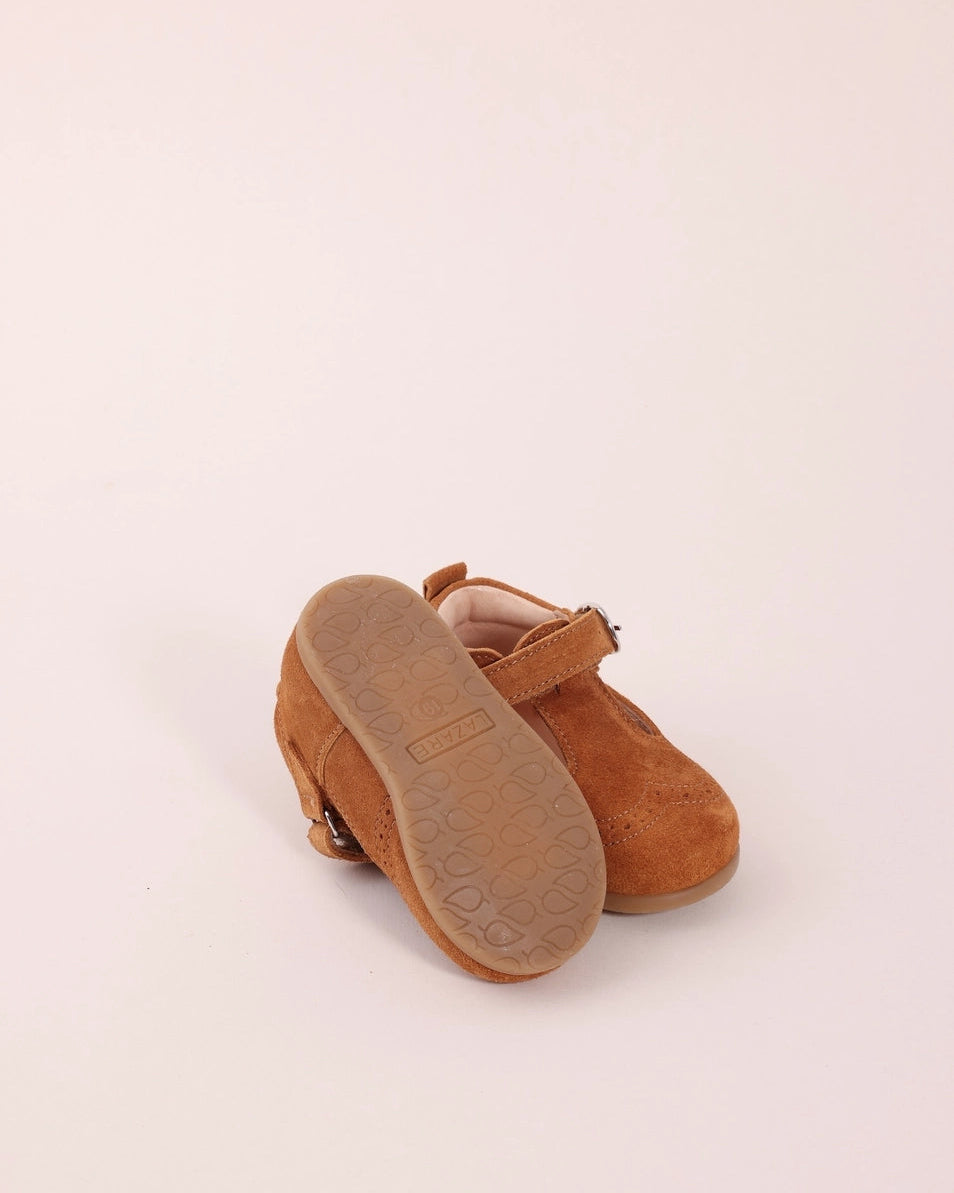 A pair of toddler Mary Jane shoes in light brown suede, shown from a rear three-quarter angle with the sole of one shoe raised to display the tread.