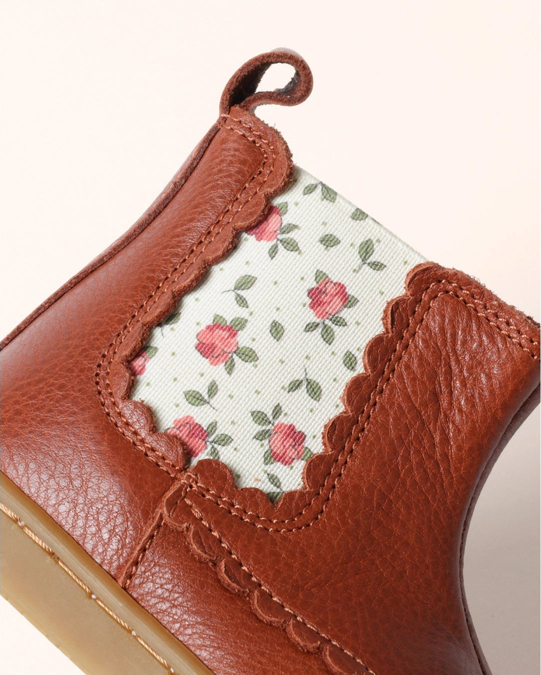 Close-up of a brown leather ankle boot with a decorative floral fabric panel and scalloped edge on a light beige background.