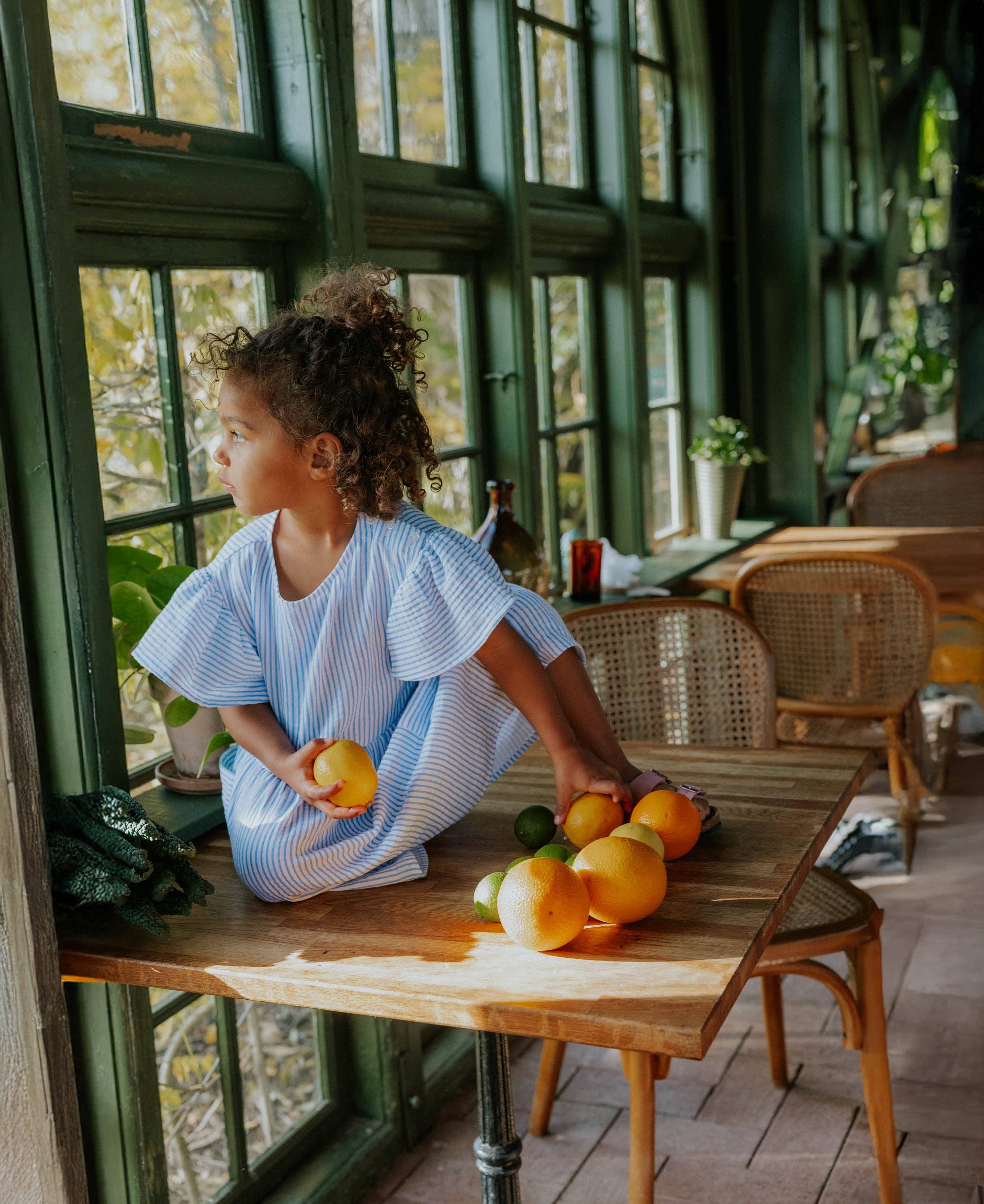 Child sitting at a table with fruits, looking out a window in a cozy room.