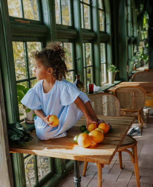 Child sitting at a table with fruits, looking out a window in a cozy room.