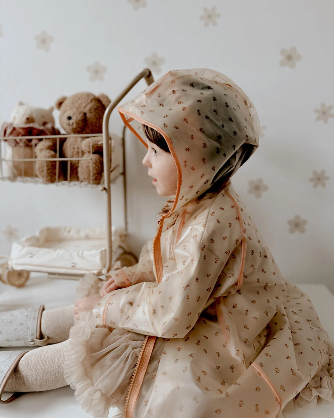 A child wearing a hooded raincoat with a small floral pattern, sitting in a softly styled room with teddy bears and a floral wall background.