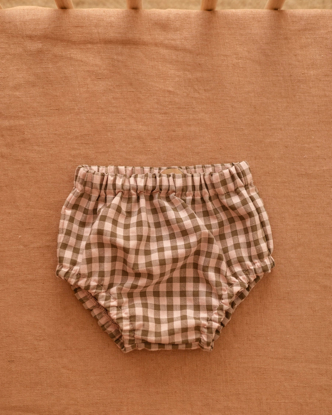 A pair of baby bloomers with a brown and beige gingham check pattern, laid flat on a textured tan linen fabric background.