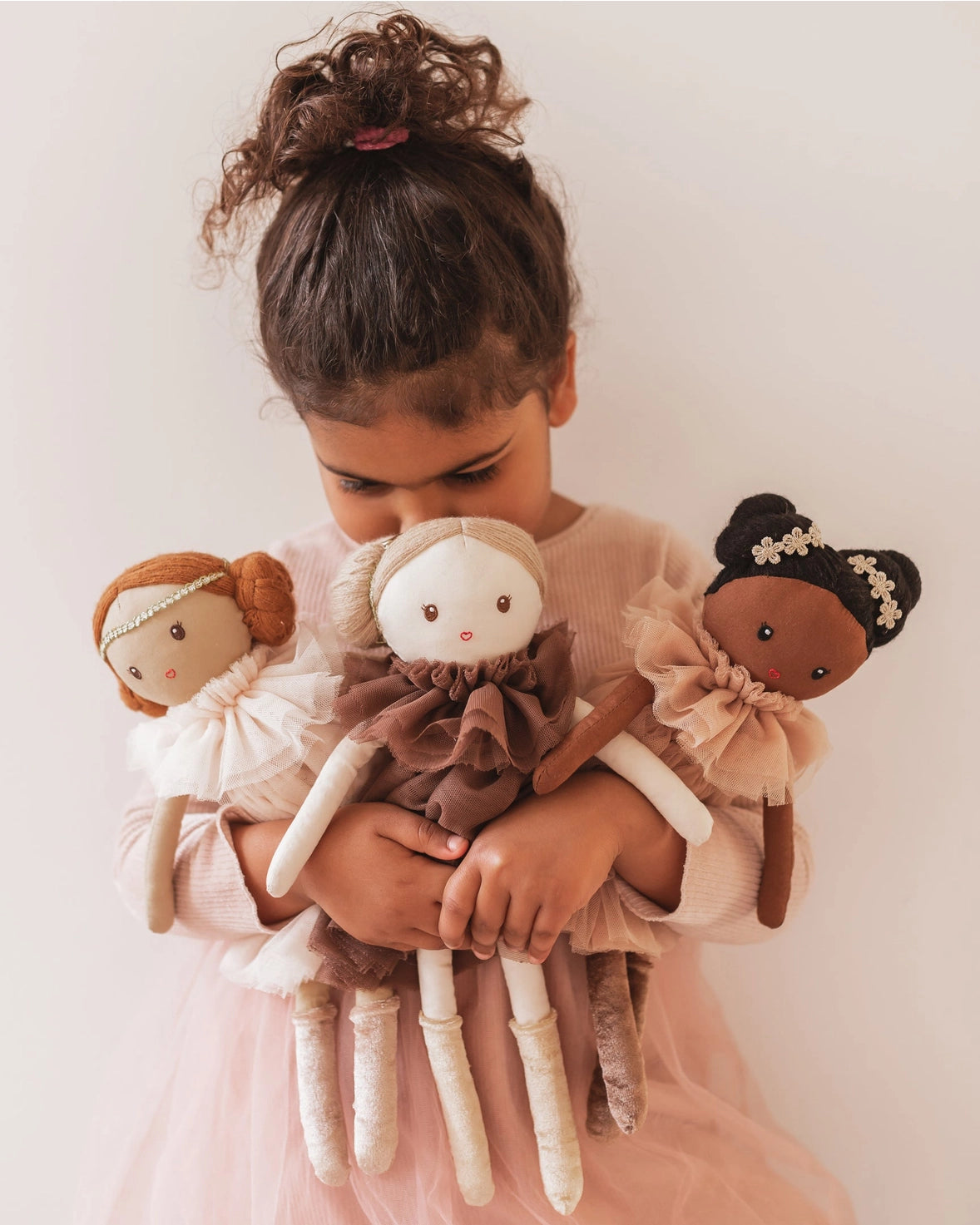 Child holding three dolls with different hairstyles and outfits against a plain background