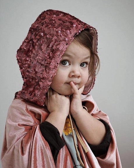 A toddler wearing a dusty rose pink sequined bonnet and a matching satin cape, posing with their fingers to their chin against a plain gray studio background.