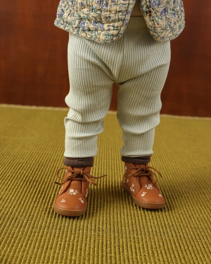 A close-up of a child's lower body wearing light sage green ribbed knit pants, brown leather lace-up shoes with white floral embroidery, and a floral quilted jacket, standing on a textured olive-green rug against a brown wooden background.
