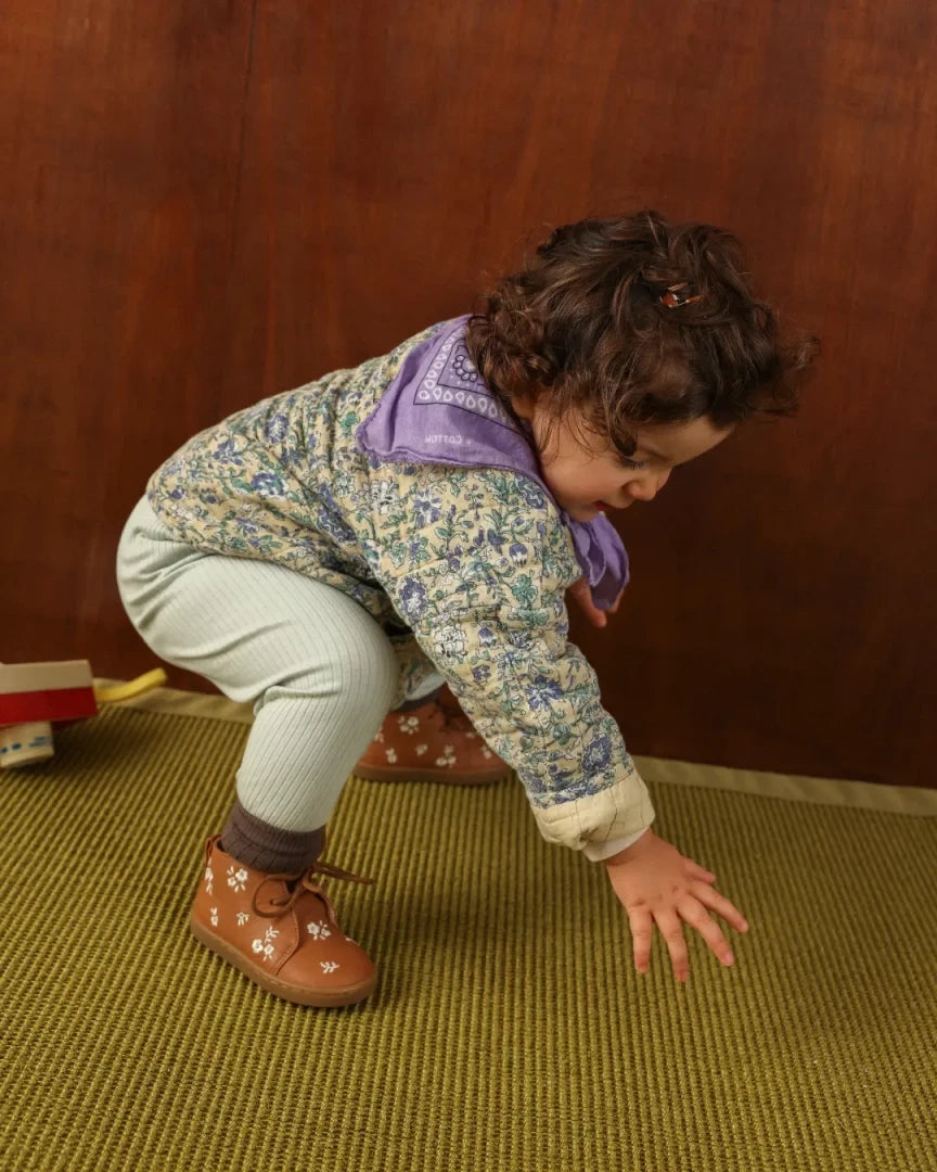 A toddler crouches on a textured olive-green rug against a dark wood wall, wearing a floral-patterned jacket with a purple hood, light green ribbed pants, and brown shoes with white floral prints.
