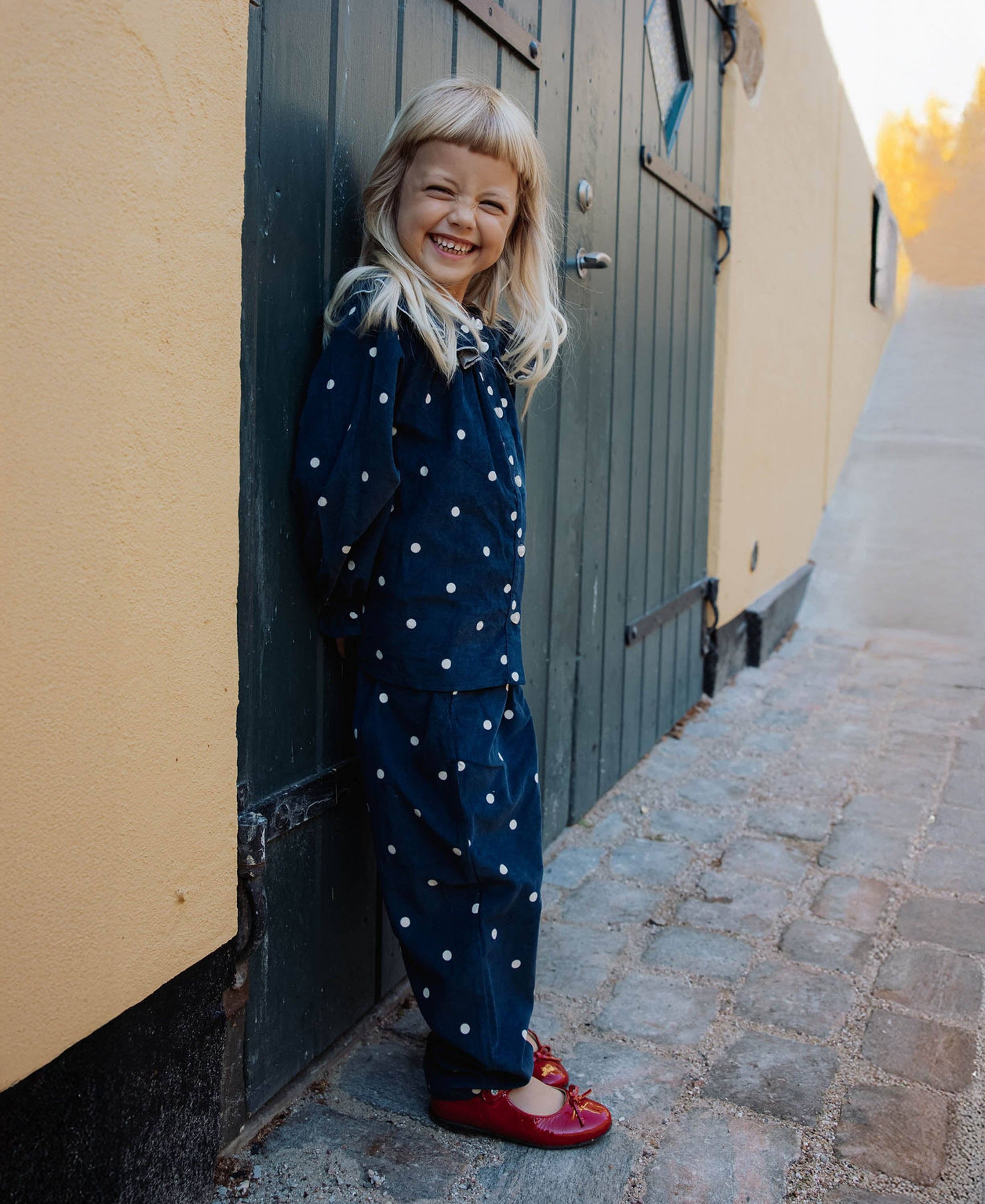 A young blonde girl smiling while leaning against a dark green wooden door on a cobblestone street. She is wearing a navy blue jumpsuit with white polka dots and red shoes.
