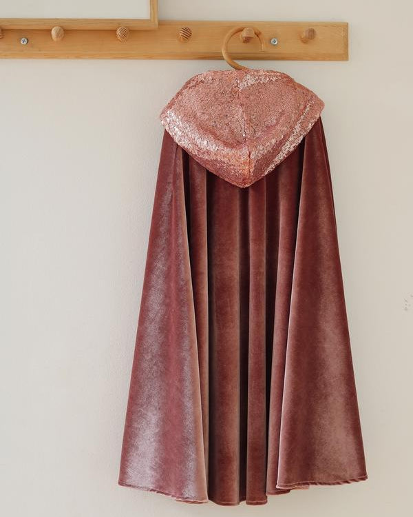 A dusty rose velvet cape with a textured, glittery rose-gold hood, hanging on a wooden hanger from a light wood peg rail against a plain off-white wall.