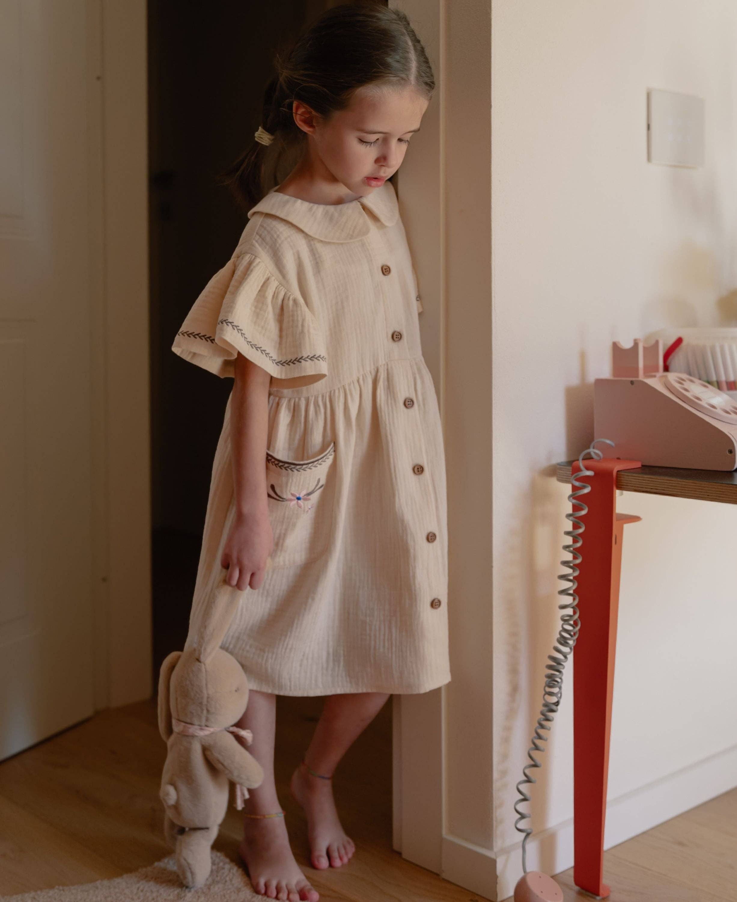 Young girl in a beige dress holding a plush toy in a room with a pink chair and white wall.