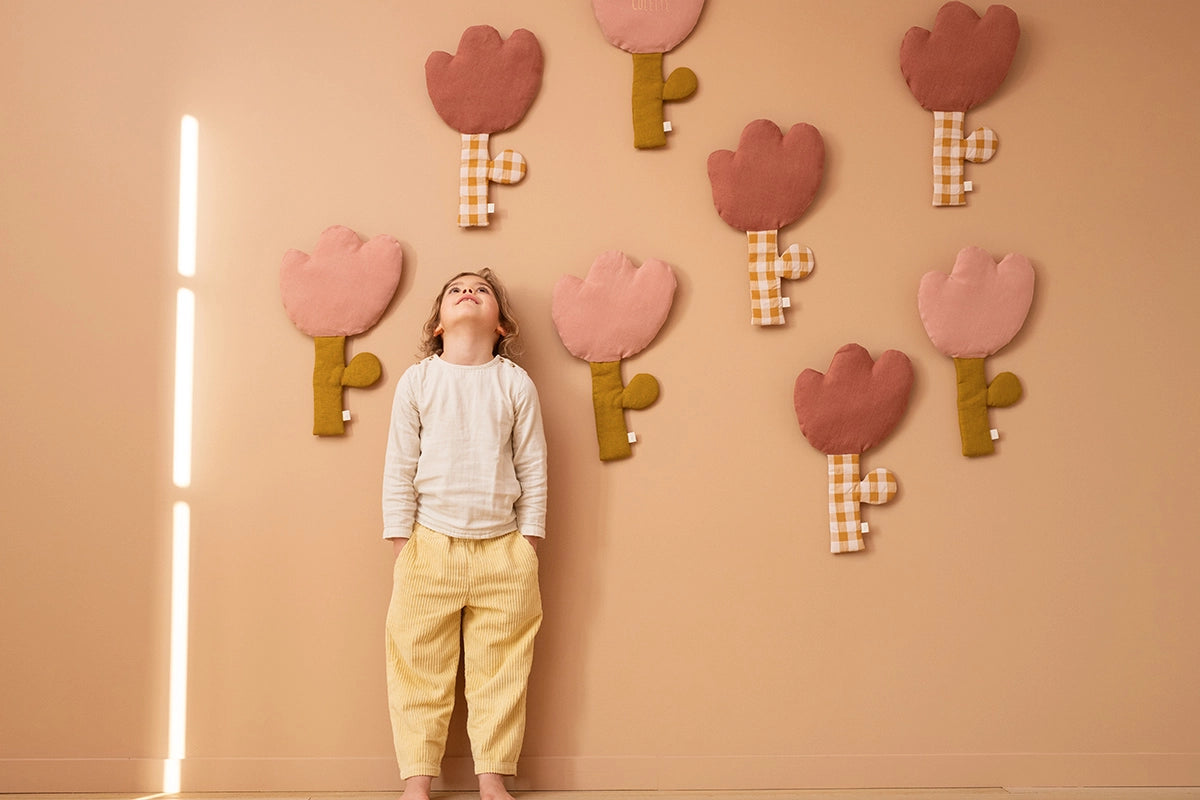 A young child stands against a peach-colored wall looking up at several decorative felt flower shapes in pink, mauve, and olive green with plaid stems.