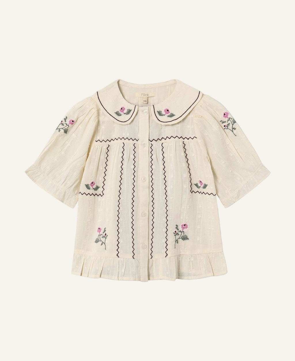 An off-white short-sleeve blouse with pink floral embroidery and black zigzag stitching details, featuring a Peter Pan collar and button-up front, shown on a light gray background.