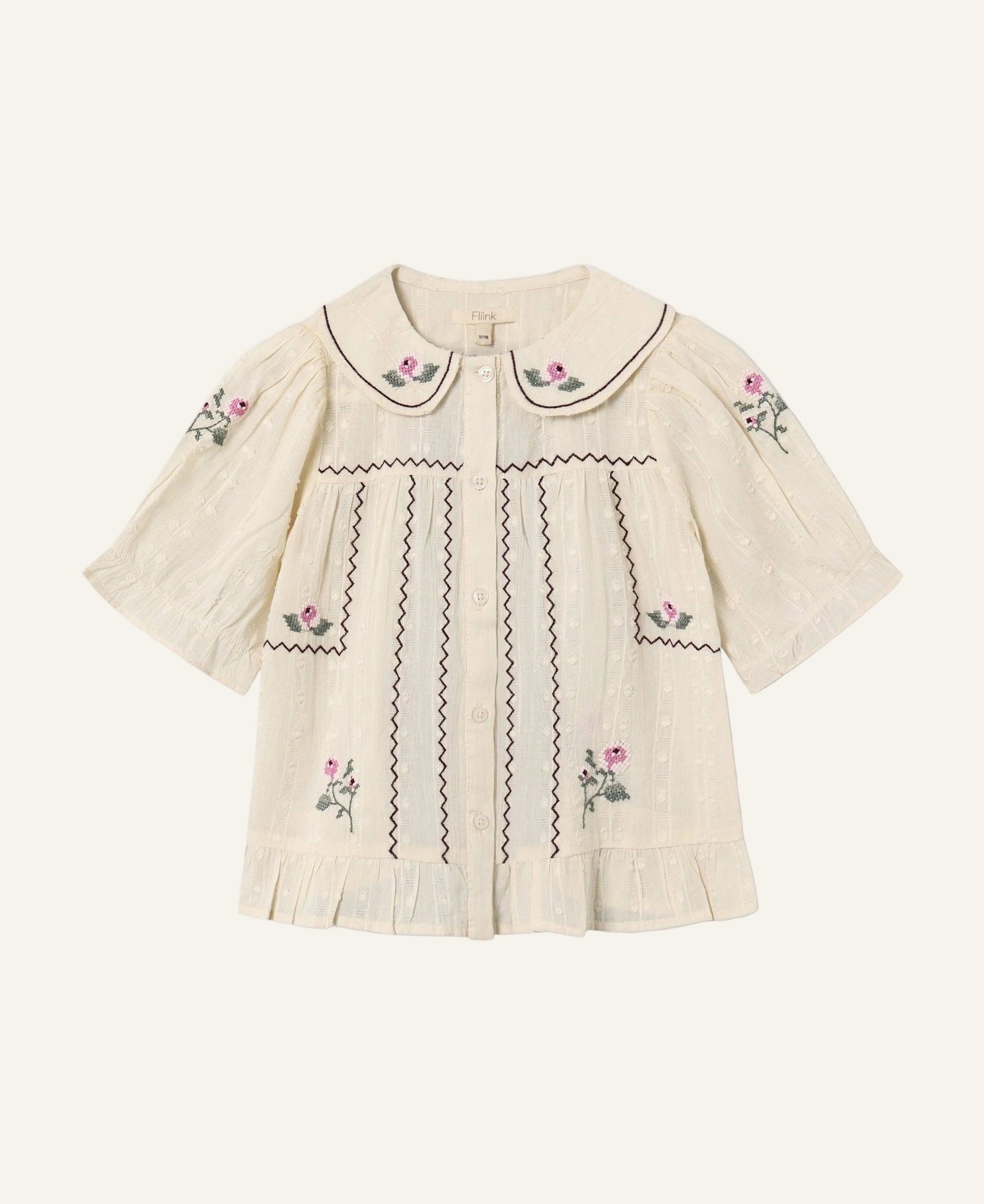 An off-white short-sleeve blouse with pink floral embroidery and black zigzag stitching details, featuring a Peter Pan collar and button-up front, shown on a light gray background.
