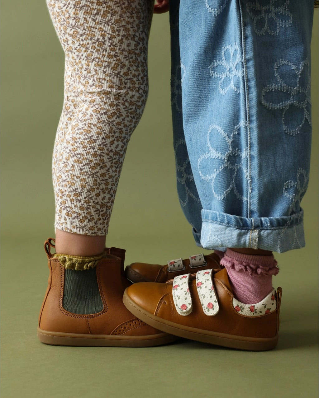 A close-up of a child's feet wearing brown suede shoes with floral trim and velcro straps, paired with patterned leggings and blue jeans, against a solid olive green background.