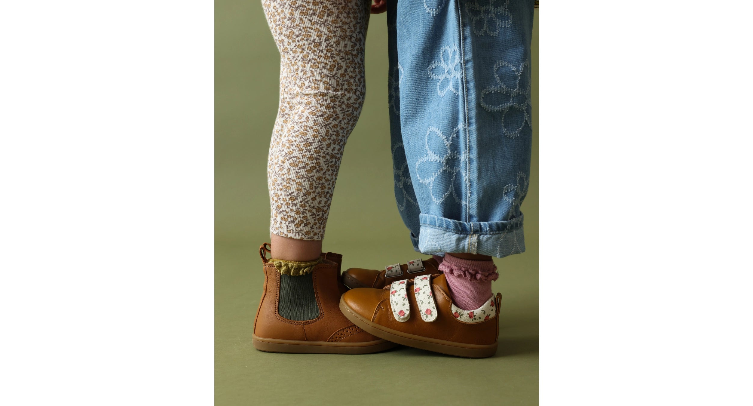 A close-up of a child's feet wearing brown suede shoes with floral trim and velcro straps, paired with patterned leggings and blue jeans, against a solid olive green background.