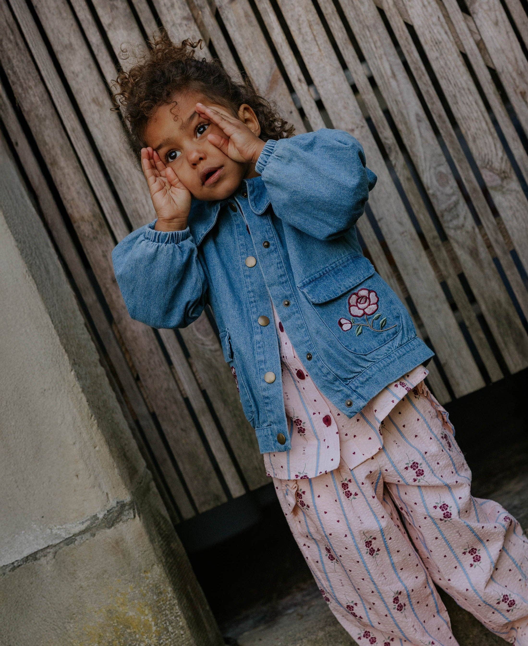 A toddler girl wearing a light blue denim jacket with a pink flower patch and a pink ruffled top with floral print, posing against a rustic wooden slat wall.