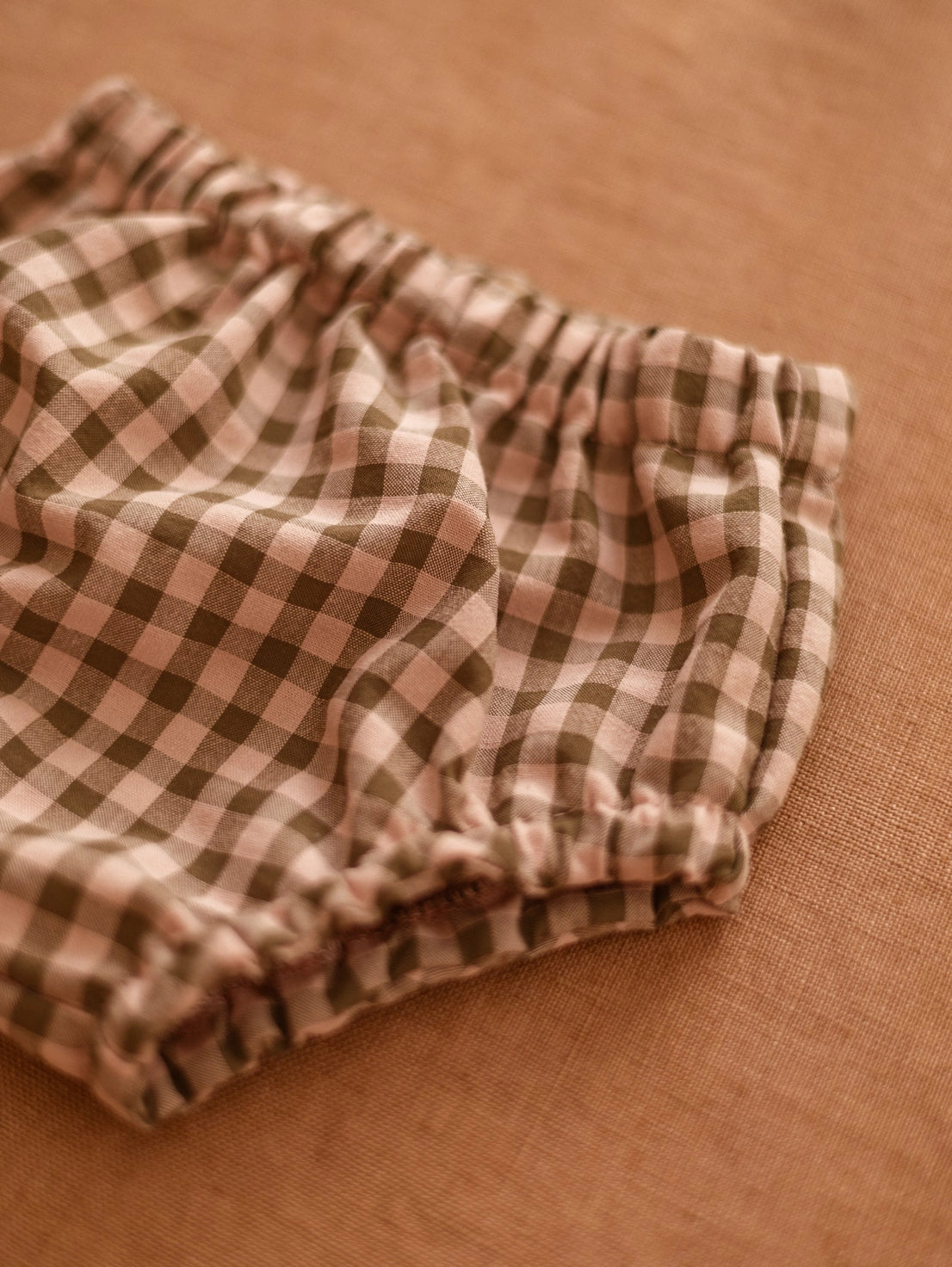 A close-up of a pair of baby bloomers with a brown and beige gingham check pattern, laid flat on a textured tan fabric background.