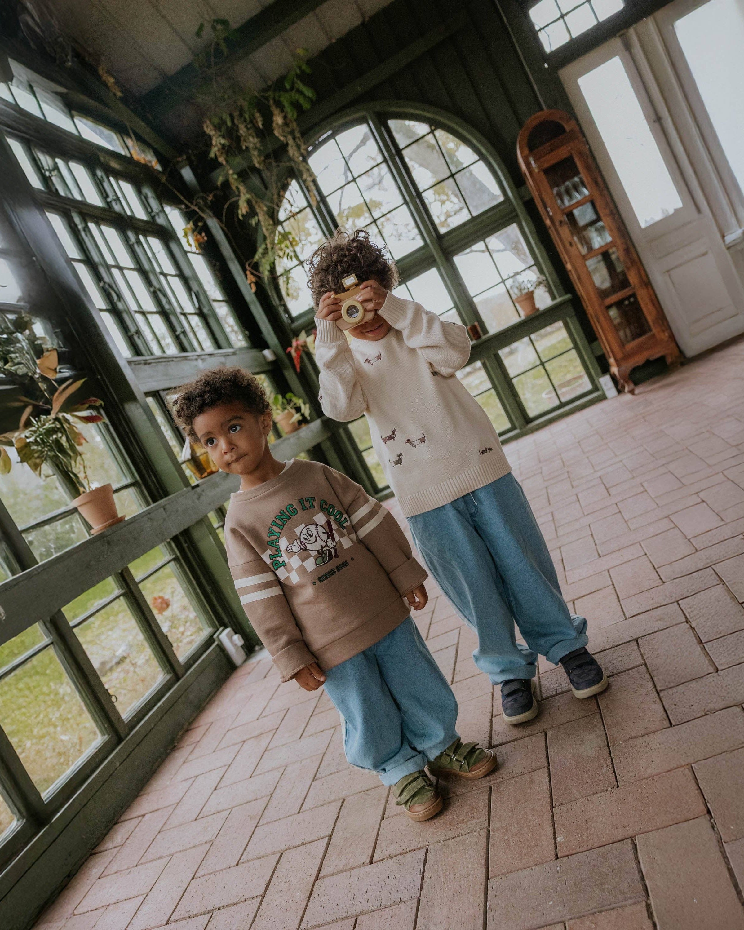 Two young boys standing on a brick porch in front of large green-framed windows. The boy on the left wears a brown graphic sweatshirt and light blue sweatpants, while the boy on the right wears a cream sweatshirt with small animal prints and light blue sweatpants.