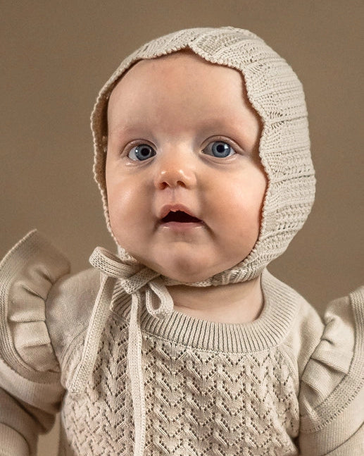 Baby wearing a beige knitted bonnet and sweater against a brown background