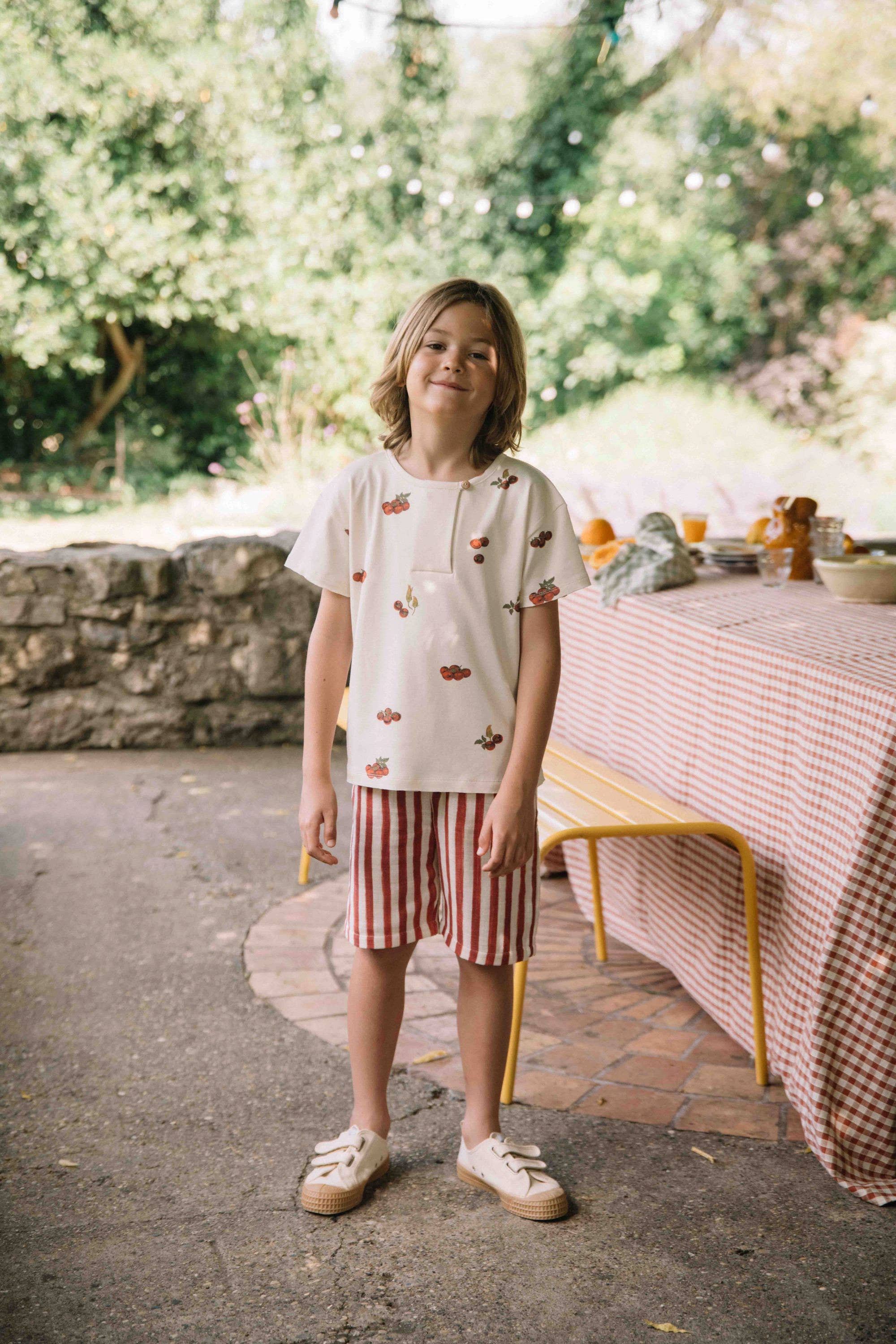 Child standing outdoors near a table with a checkered tablecloth, wearing a white shirt with red patterns and striped shorts.