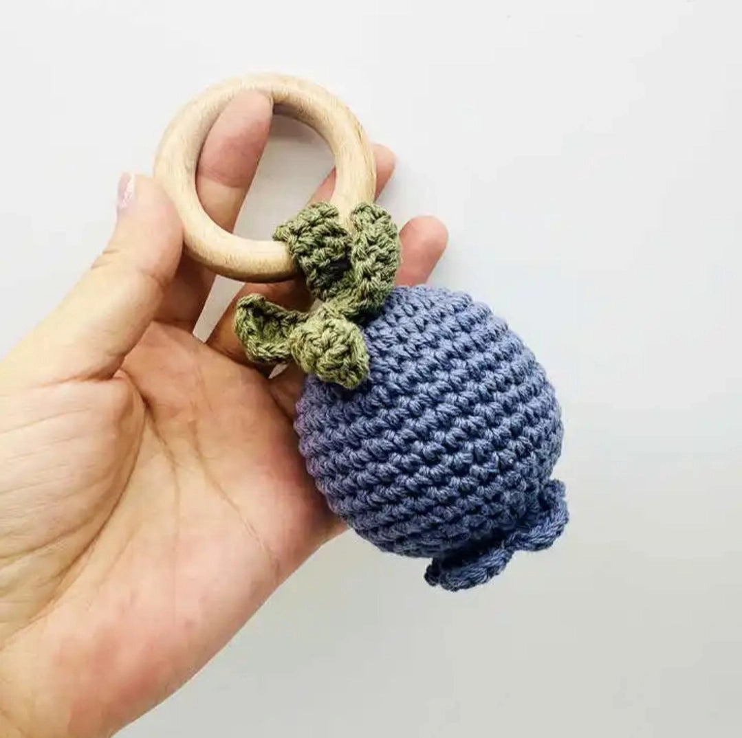 Hand holding a wooden teething ring with a blue knitted ball and green leaves on a white background
