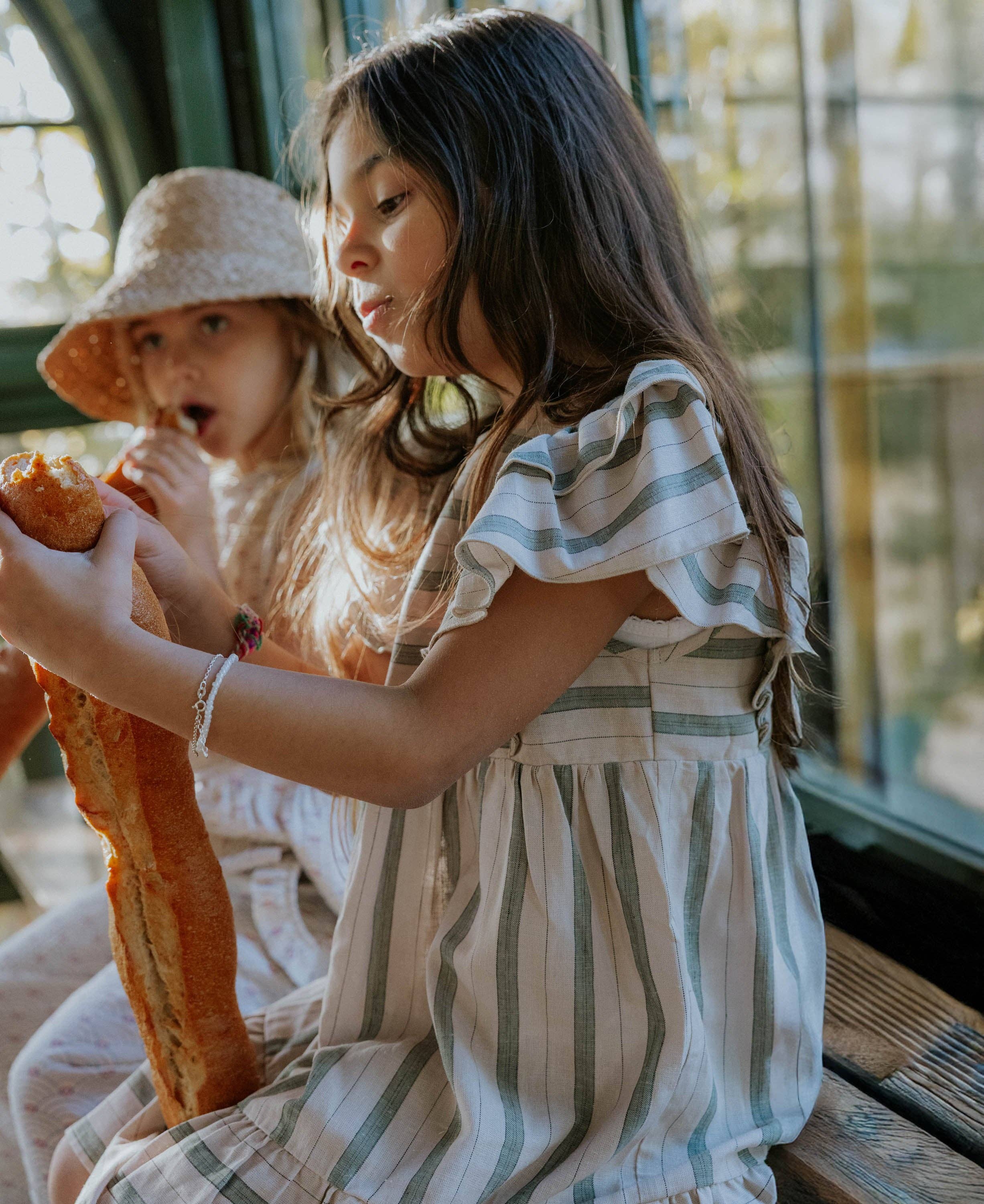 Two children sitting together, one eating a sandwich, with a blurred background.