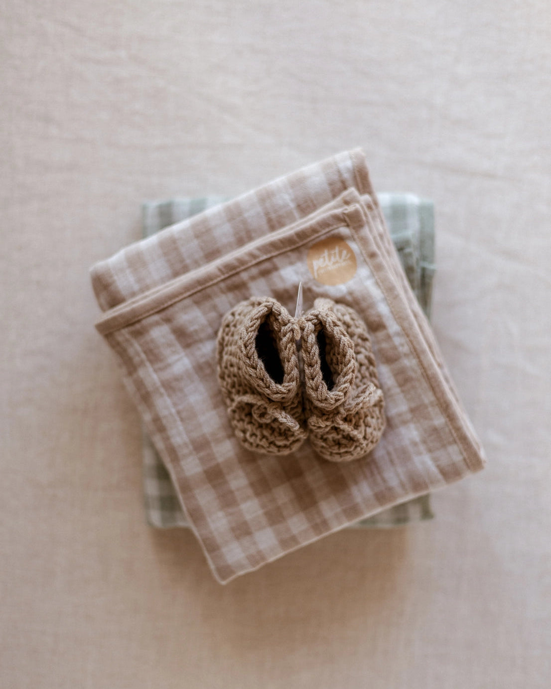 A pair of beige hand-knit baby booties resting on a folded beige and white gingham blanket, which is stacked on top of a green plaid cloth against a neutral beige background.