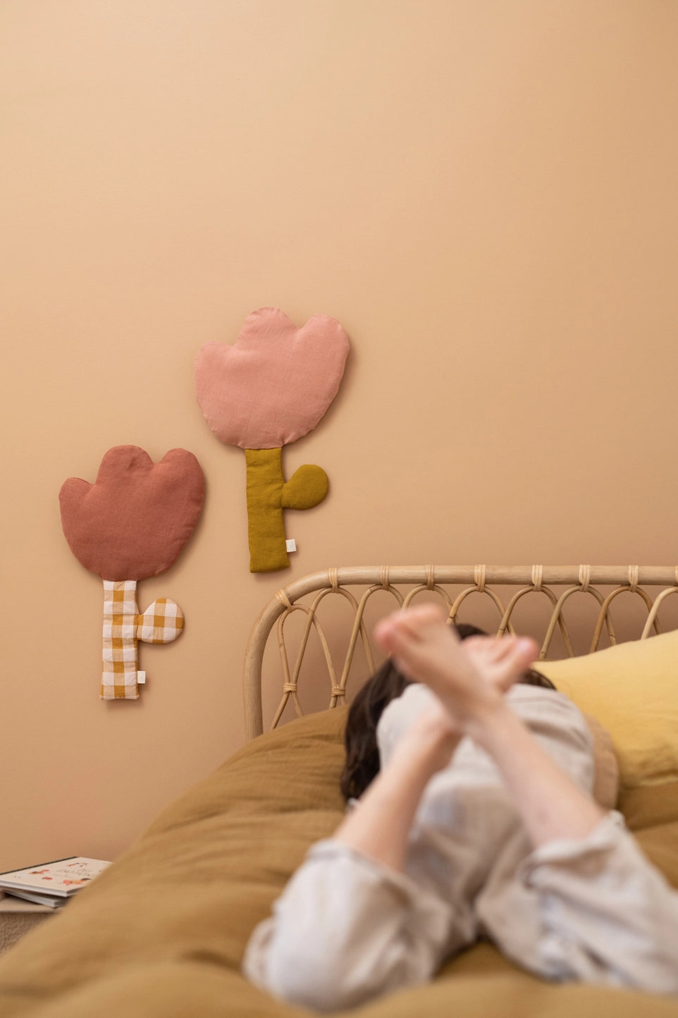 A person lying on a bed in a cozy bedroom, with two decorative tulip-shaped wall hangings in pink and mauve on the beige wall above the bed.