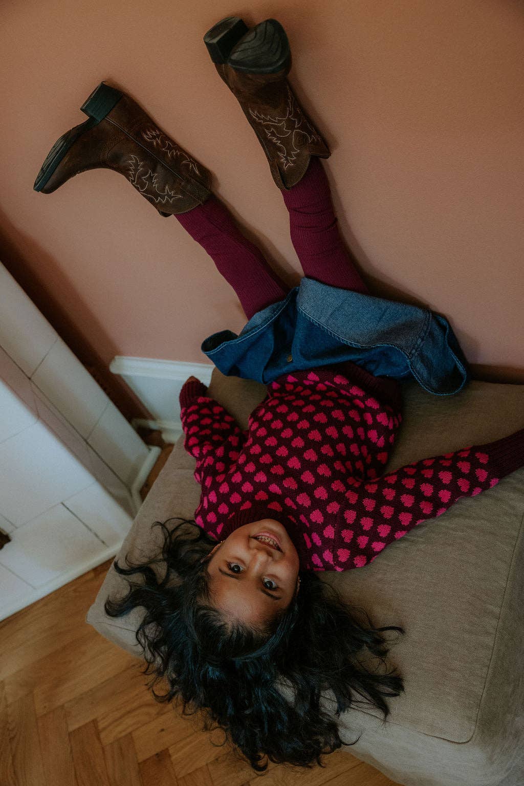 A child lying on a beige couch in a home setting, wearing a pink and black patterned long-sleeve top, purple leggings, a blue denim skirt, and brown cowboy boots against a dusty pink wall.