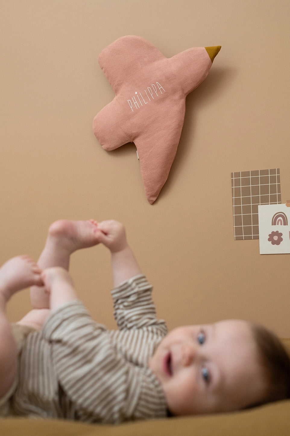 A baby wearing a beige and white striped romper lies on their back on a tan surface, looking up at a pink bird-shaped wall hanging with the name 'MARTHA' embroidered on it.