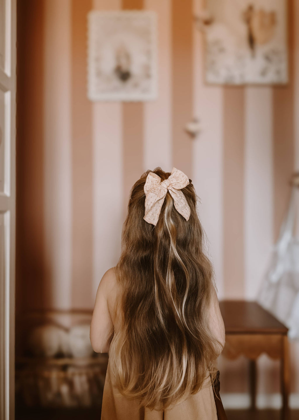 Child with a large bow in their hair standing in a room with striped wallpaper.