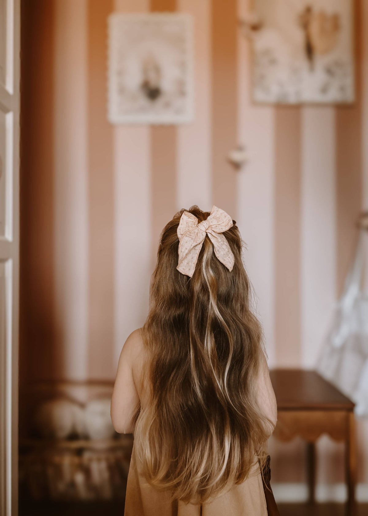 Child with a large bow in their hair standing in a room with striped wallpaper.