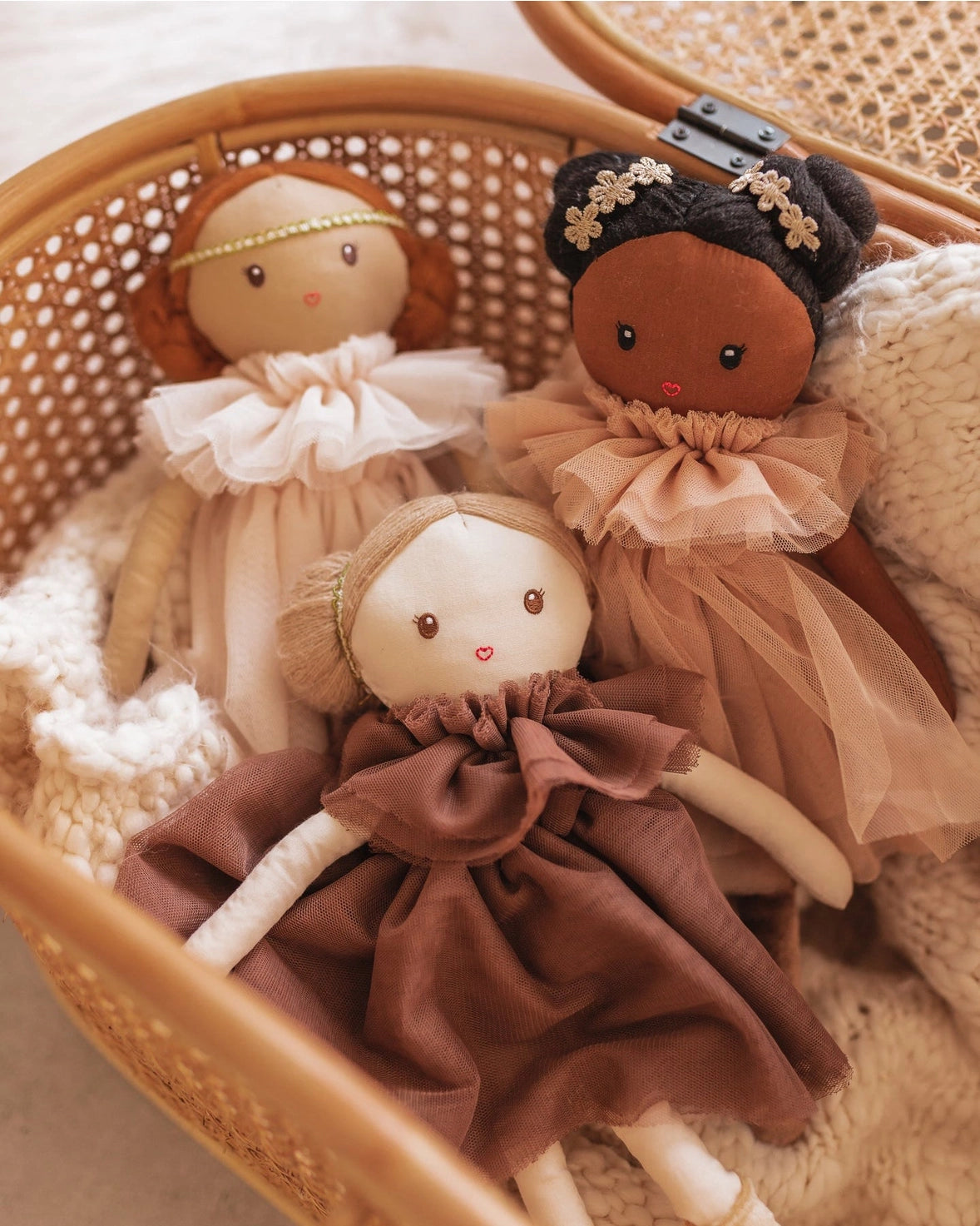 Three handmade fabric dolls with embroidered faces and ruffled dresses, arranged inside a woven rattan basket on a soft cream knit blanket.