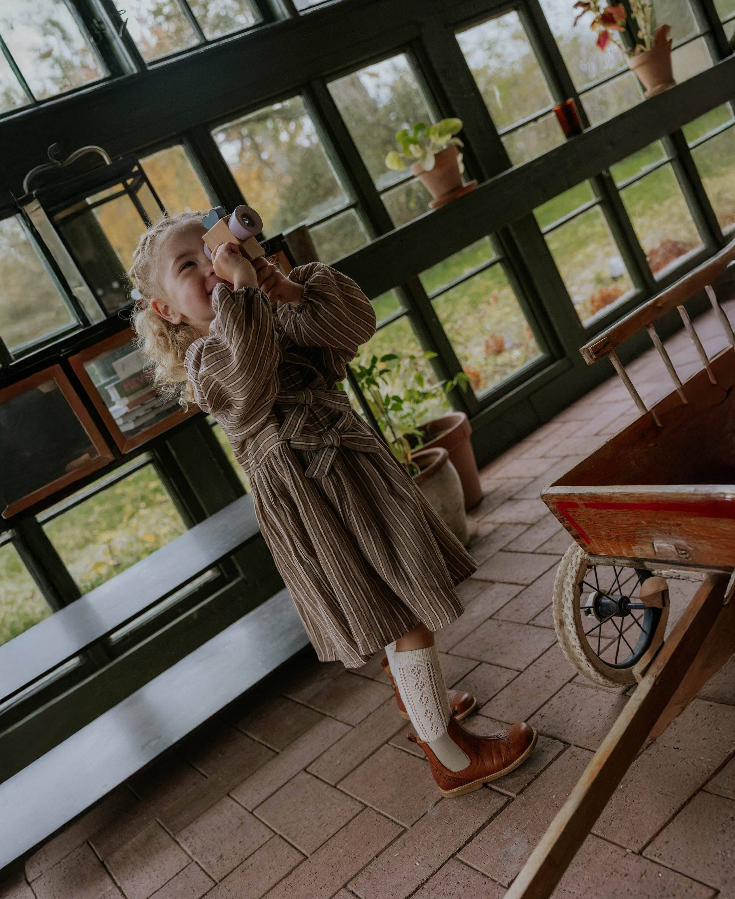 A young girl with blonde curly hair stands in a rustic greenhouse or sunroom, holding a toy camera to her eye. She is wearing a brown and beige vertically striped long-sleeve dress, white knit socks, and brown leather shoes.
