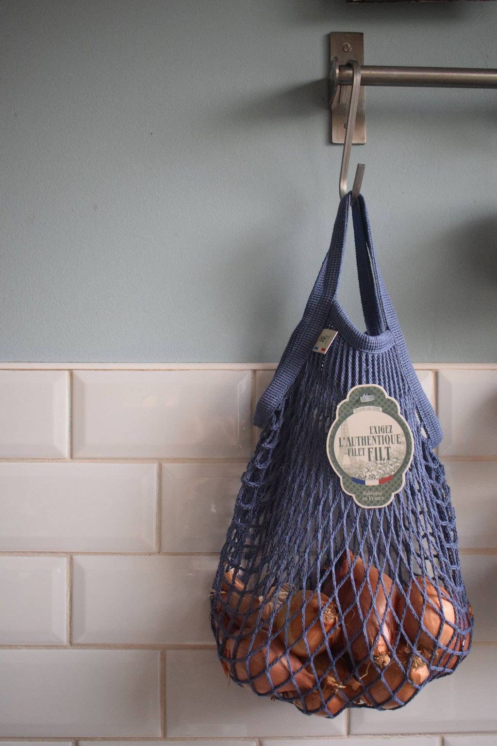 Blue mesh bag with produce hanging on a hook against a tiled wall.