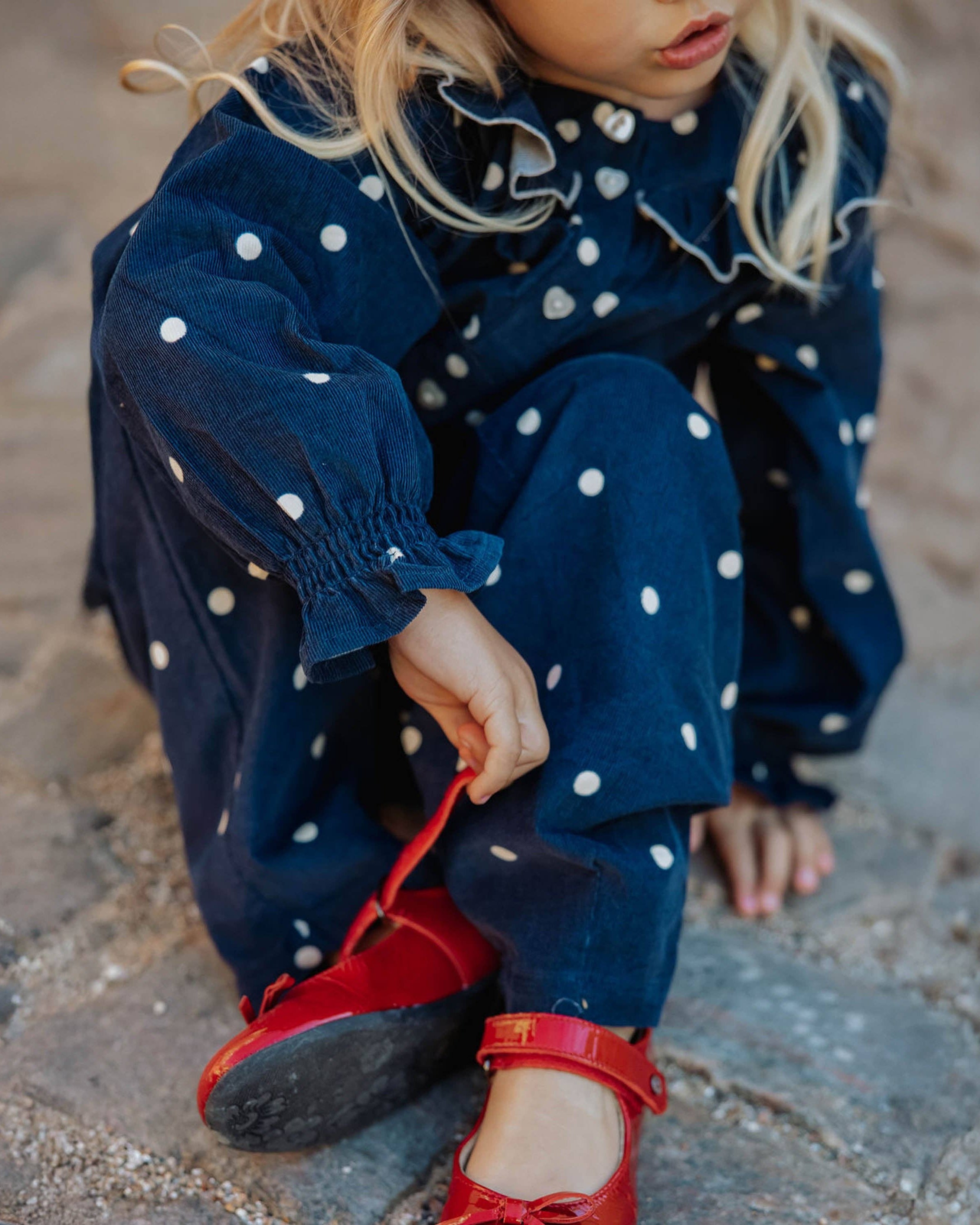 A young child wearing a navy blue corduroy jumpsuit with white polka dots, sitting on a stone surface and tying the red laces of red shoes.