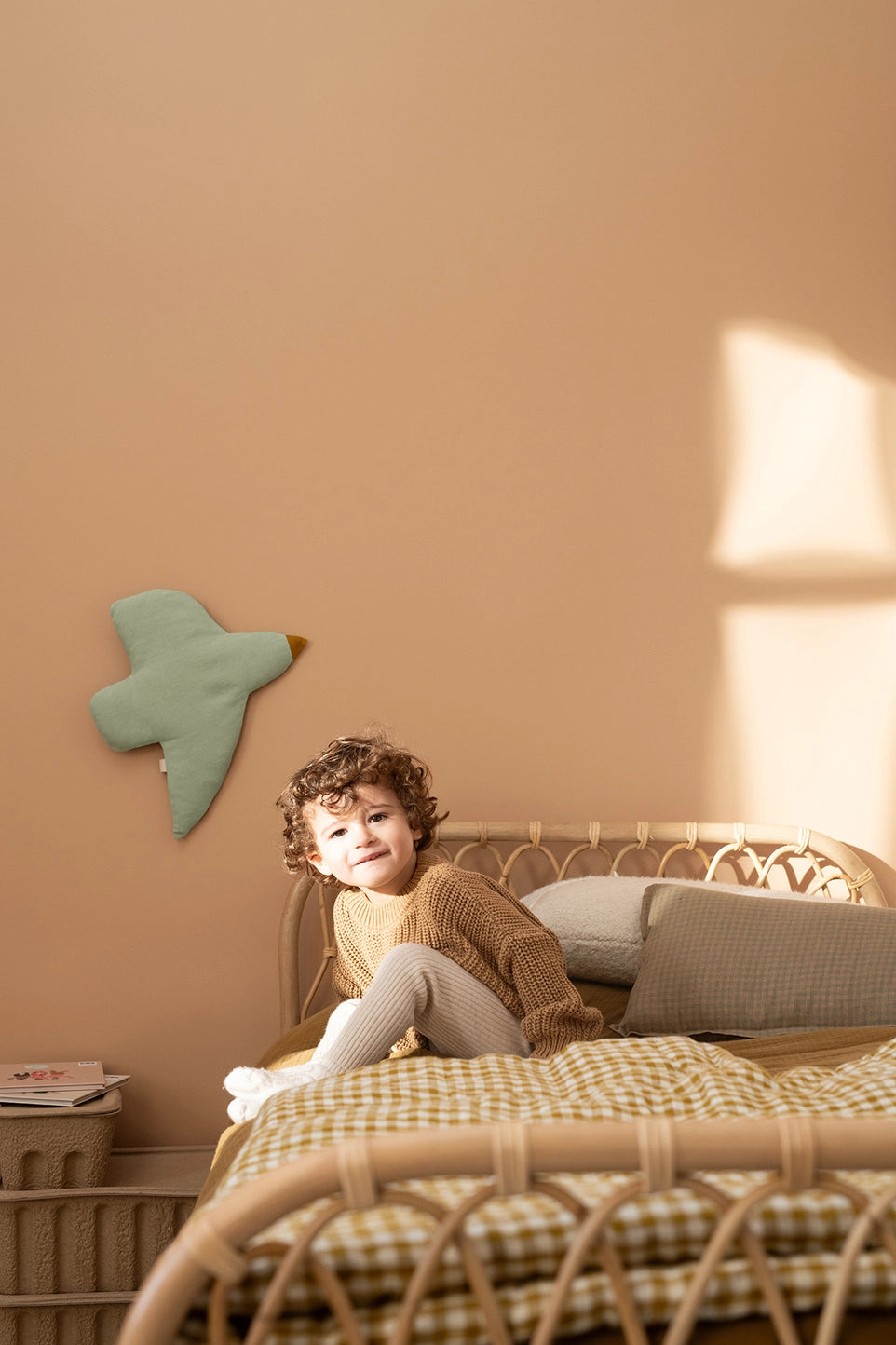 A toddler with curly brown hair sits on a rattan bed in a warm, sunlit room. The child is wearing a beige knit sweater and light gray leggings.
