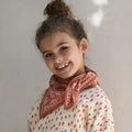 A young girl with her hair in a high bun smiles while wearing a rust-orange patterned bandana around her neck and a cream top with a red leaf print.