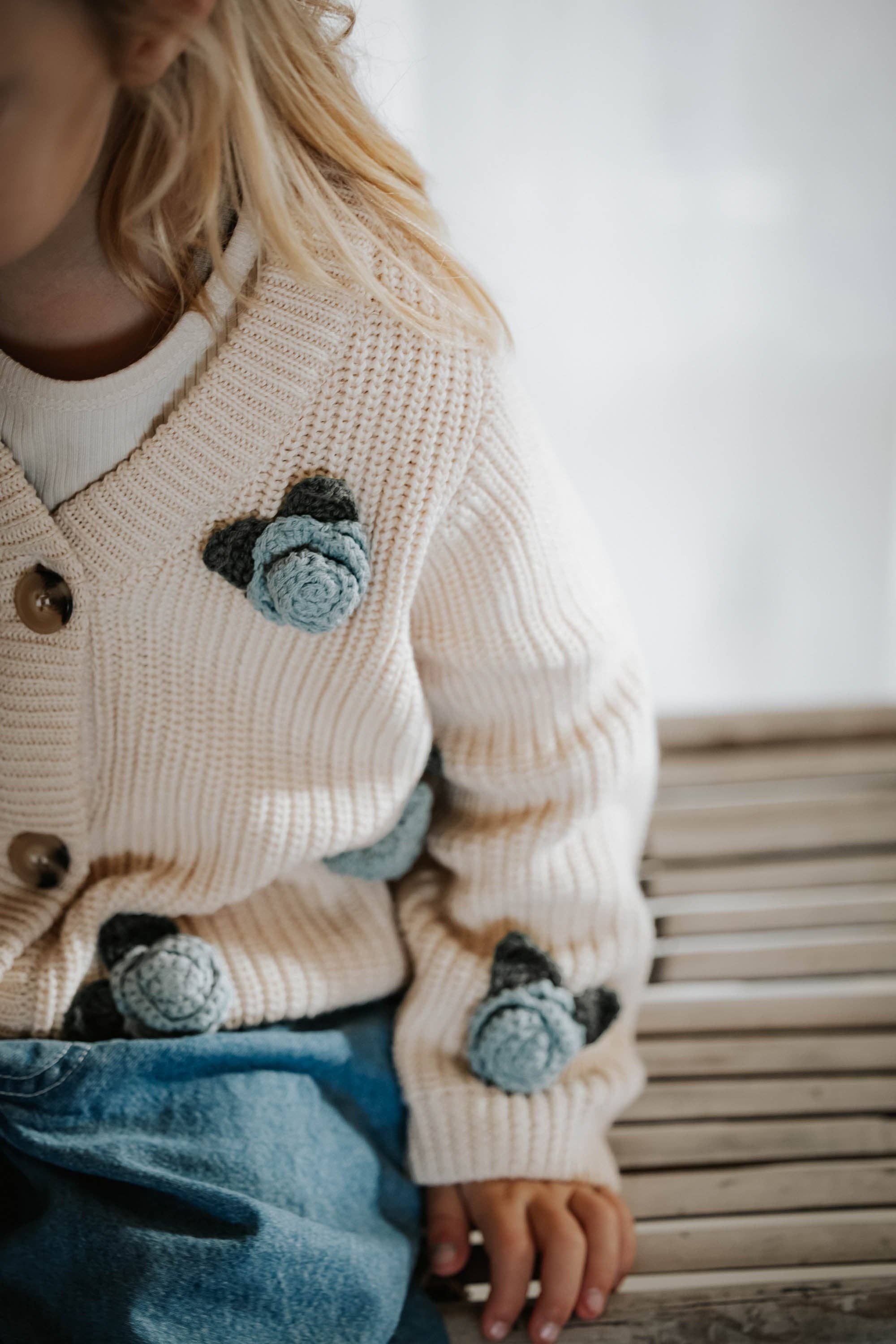 Close-up of a child wearing a cream-colored knitted cardigan with black and blue floral patterns, sitting on a wooden bench.