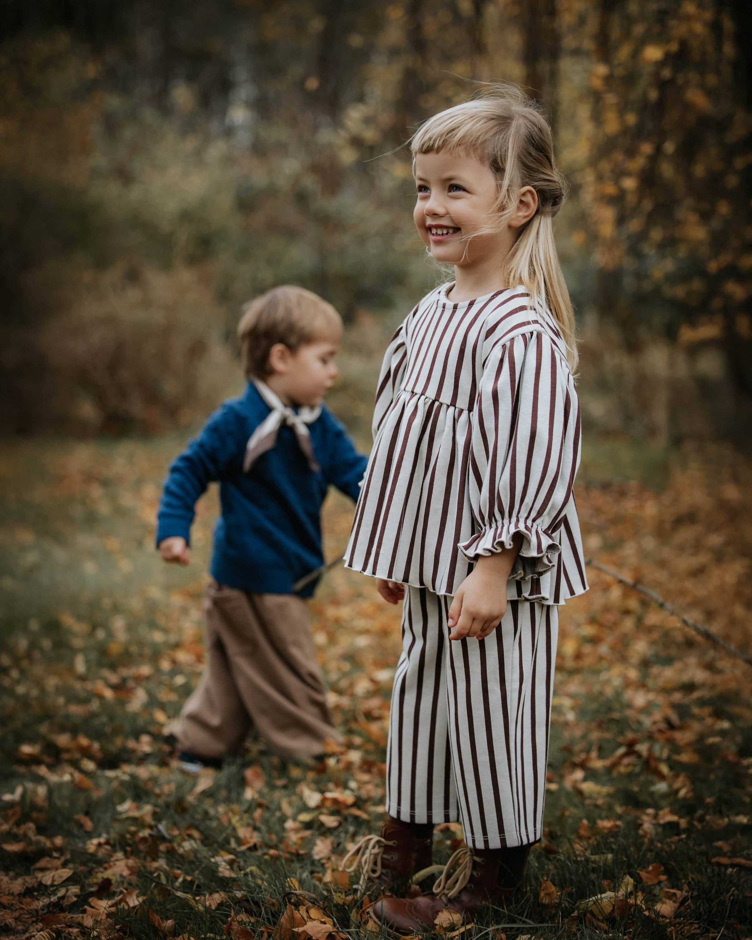 A young girl stands outdoors in a wooded, leaf-covered area wearing a matching two-piece outfit with vertical brown and cream stripes, while a young boy in a blue sweater and brown pants walks behind her.