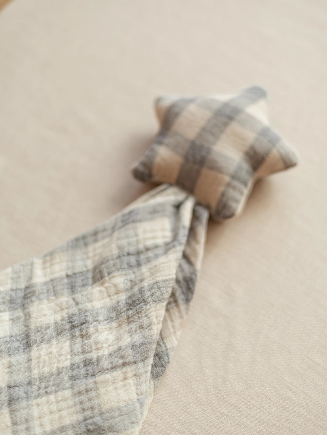 A soft plaid baby swaddle blanket in beige, cream, and gray, featuring a small matching star-shaped pillow, laid out on a neutral beige fabric background.