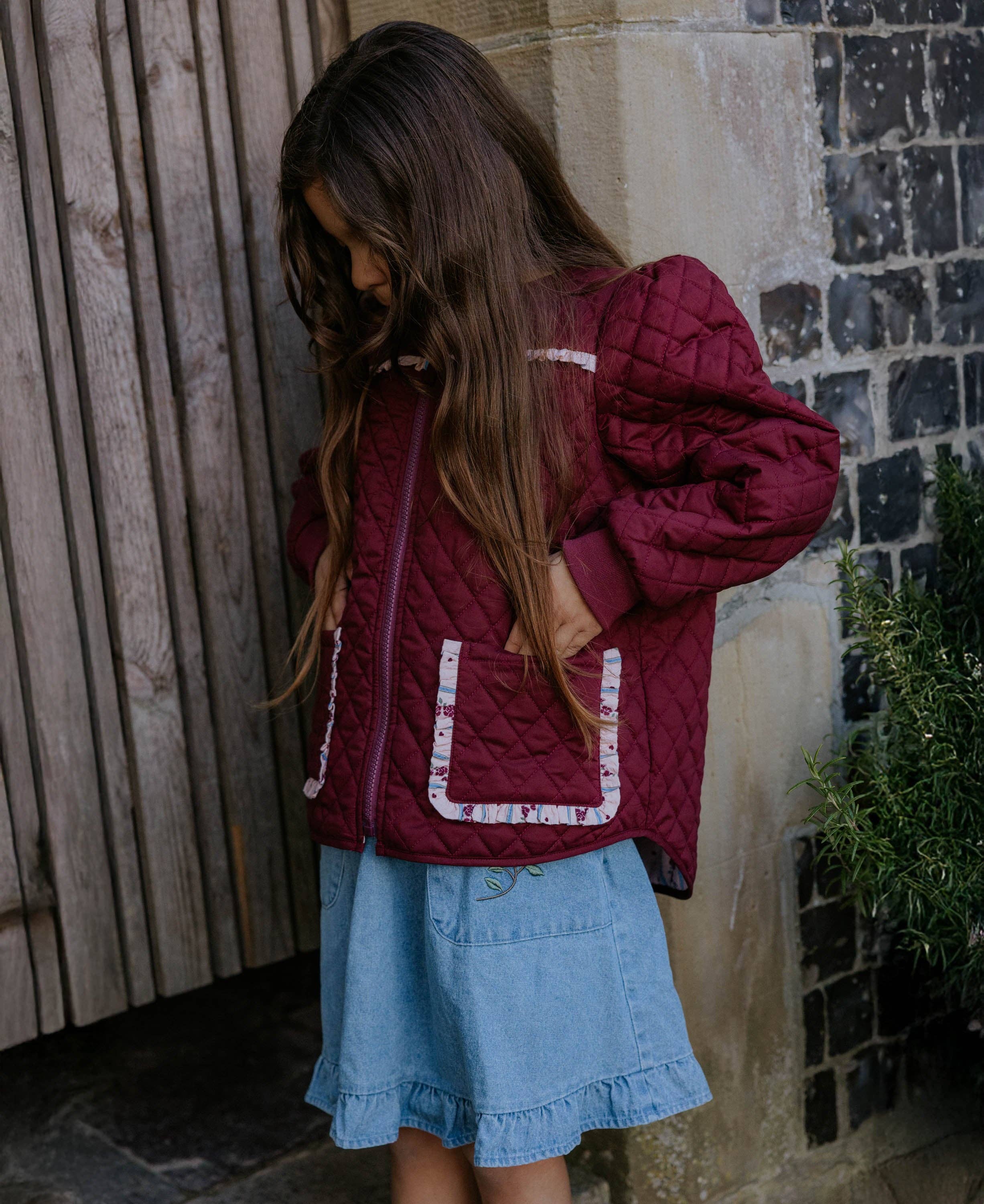 A young girl with long brown hair is shown from the side, wearing a quilted burgundy jacket with white patterned trim and a light blue denim skirt, standing outdoors against a stone wall and wooden door.
